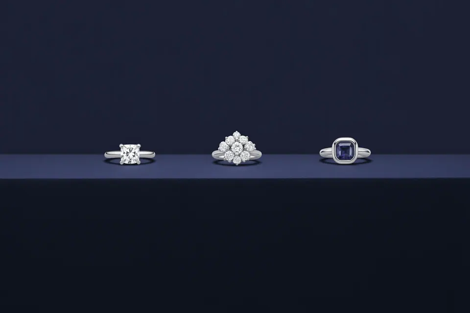 Rings with a clear central stone, a clustered design and a blue rectangular stone on a dark shelf.